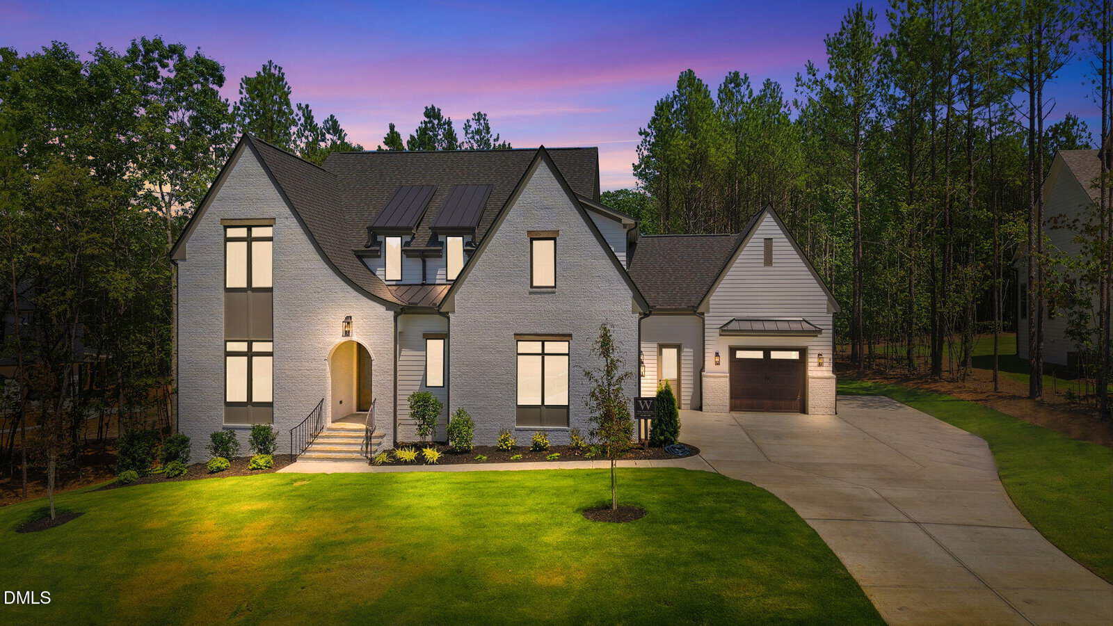 A modern two-story white brick house at 80 Spanish Oak Drive, Youngsville, features large windows, a well-lit entrance, and an attached garage. It sits on a manicured lawn surrounded by tall trees beneath a purple and blue dusk sky.