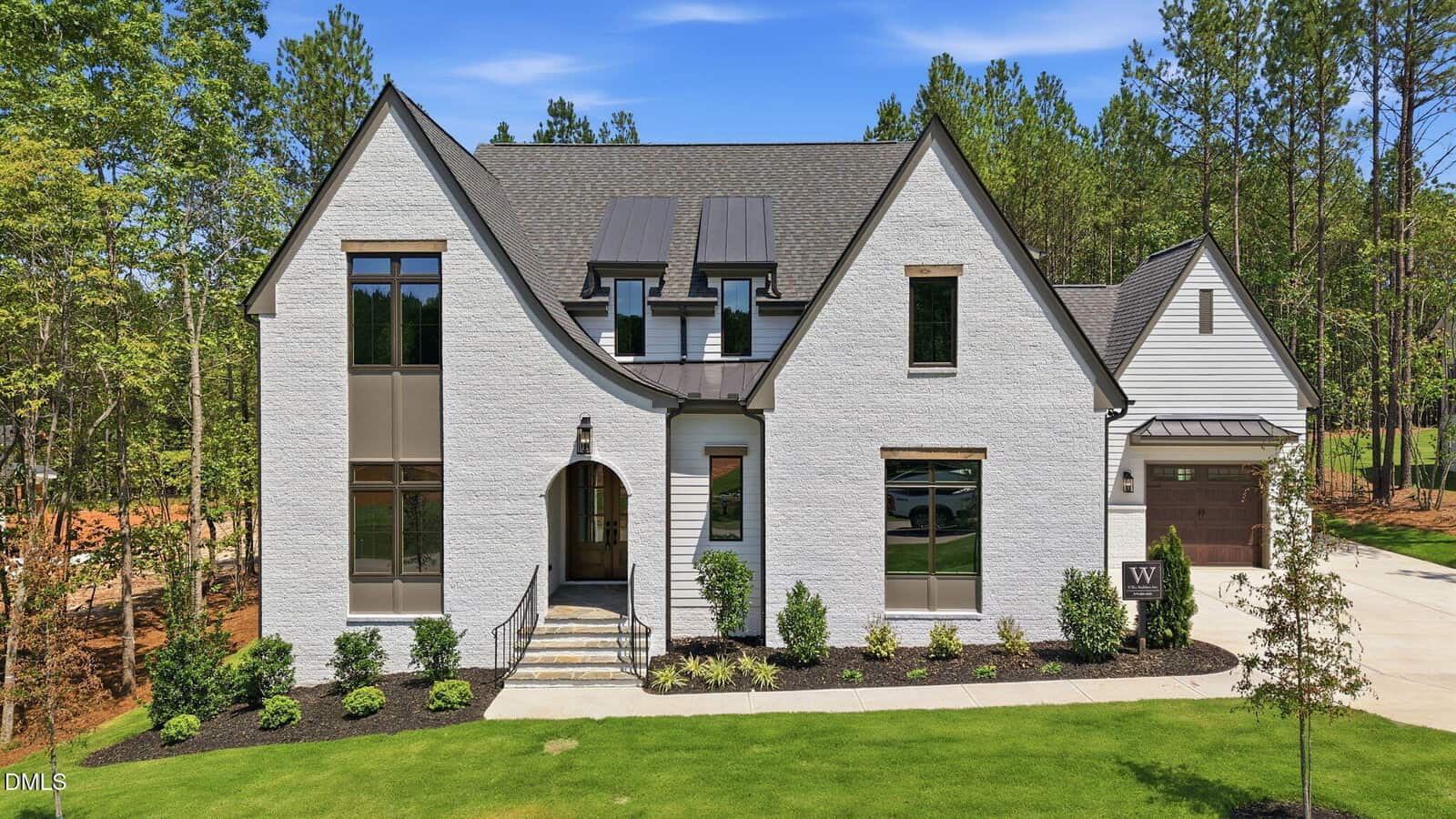 A modern two-story white brick house with large windows, dark trim, and gabled roof, surrounded by landscaped greenery and trees. A driveway leads to an attached garage on the right.