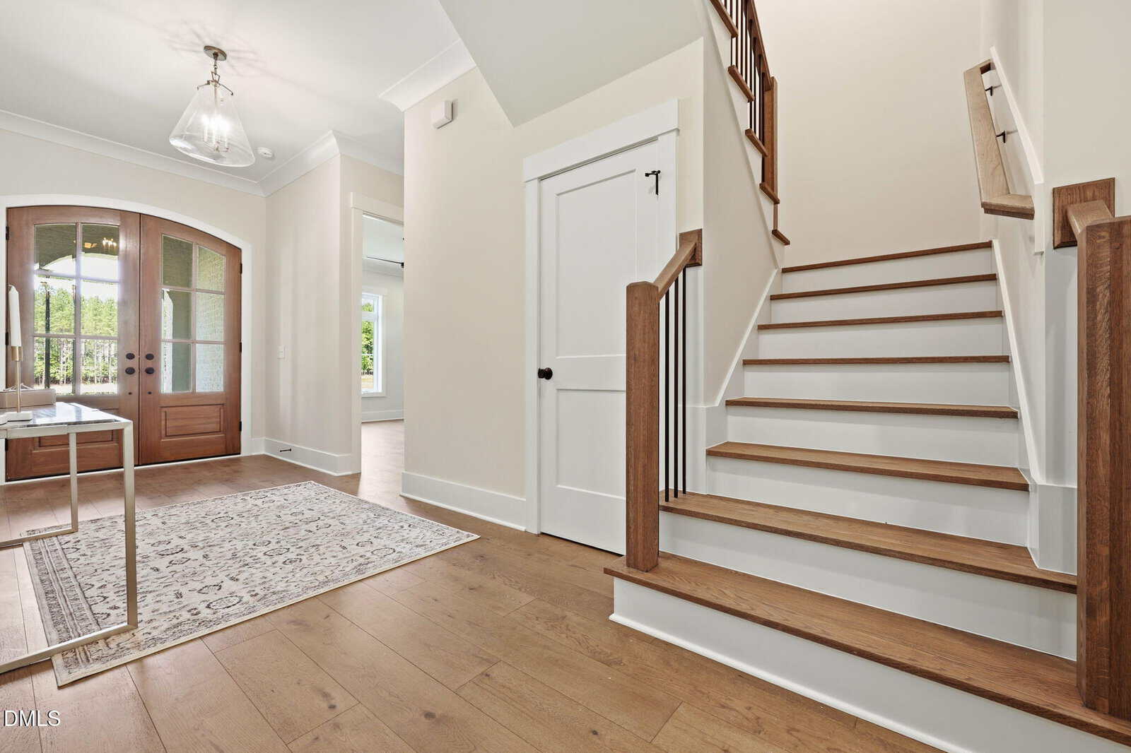 Bright entryway with wood floors, a patterned rug, a glass pendant light, a wooden front door with glass panels, a staircase with wooden steps and white risers, and light-colored walls.