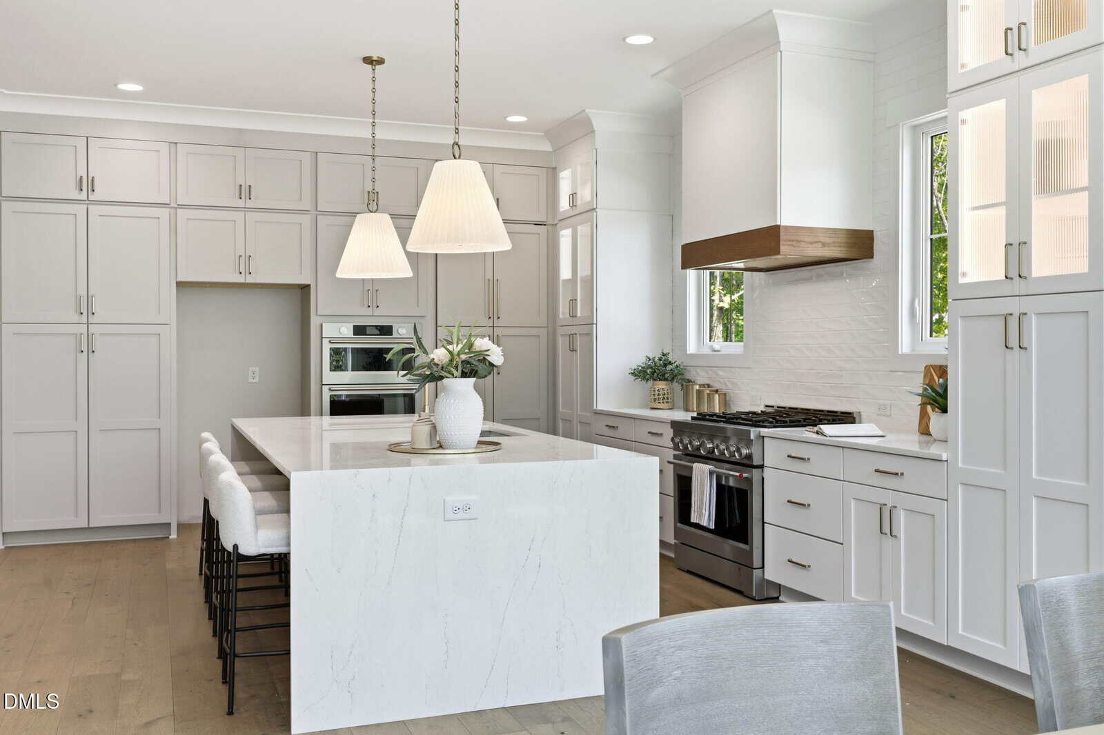 Modern white kitchen with a large marble island, four barstools, pendant lights, built-in ovens, gas stove, white cabinets, and windows letting in natural light. A vase with flowers sits on the island.