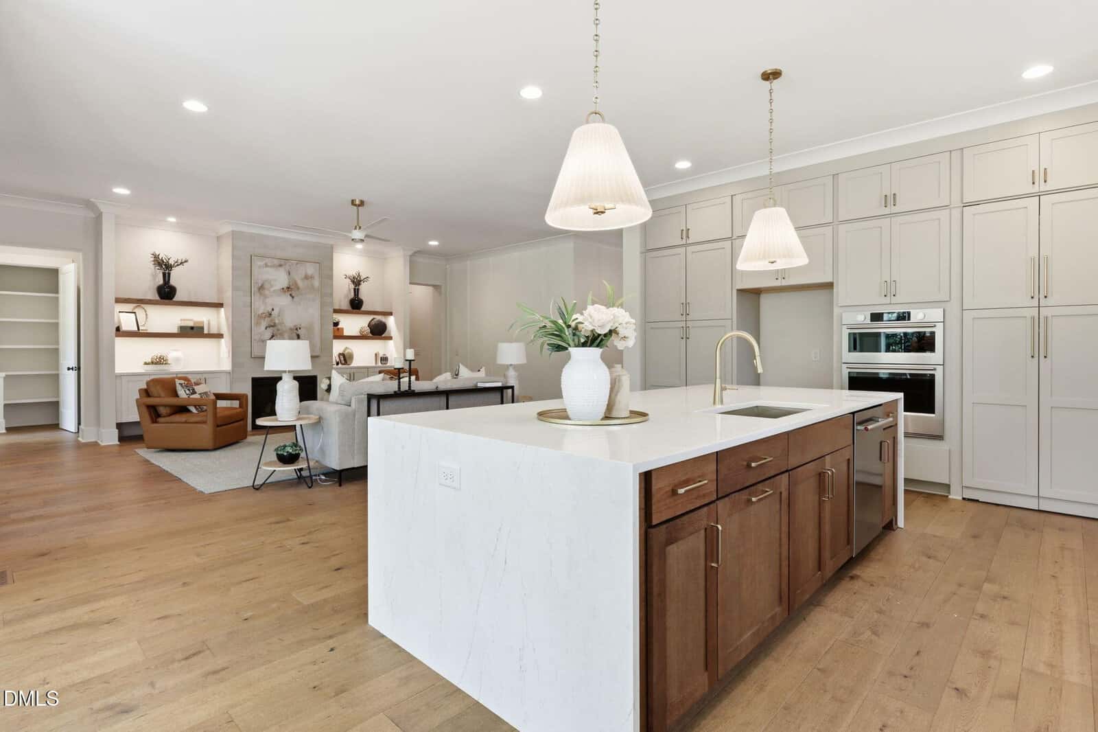 Modern open kitchen with large island, white countertops, pendant lights, built-in appliances, light wood floors, and a view into a cozy living room with shelves and neutral decor.