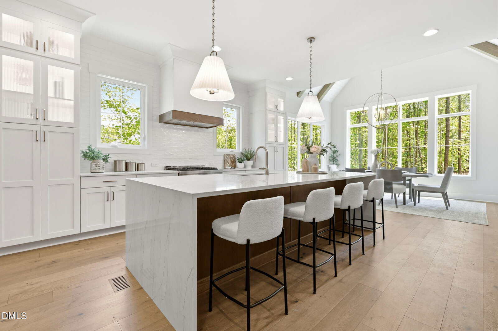 Modern kitchen with white cabinets, large island with marble countertop, four upholstered bar stools, pendant lights, and a dining area by large windows overlooking green trees. Light wood floors and bright natural light.