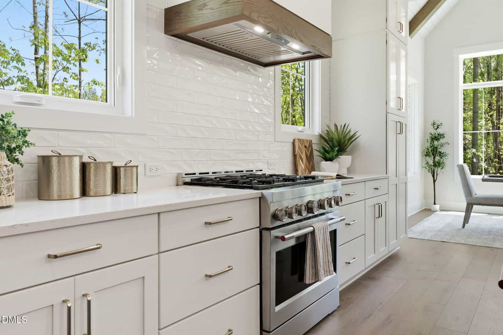 Modern white kitchen with stainless steel gas range, metal canisters, potted plant, textured white backsplash, and large windows letting in natural light. Light wood floors and minimal decor create a bright, airy feel.