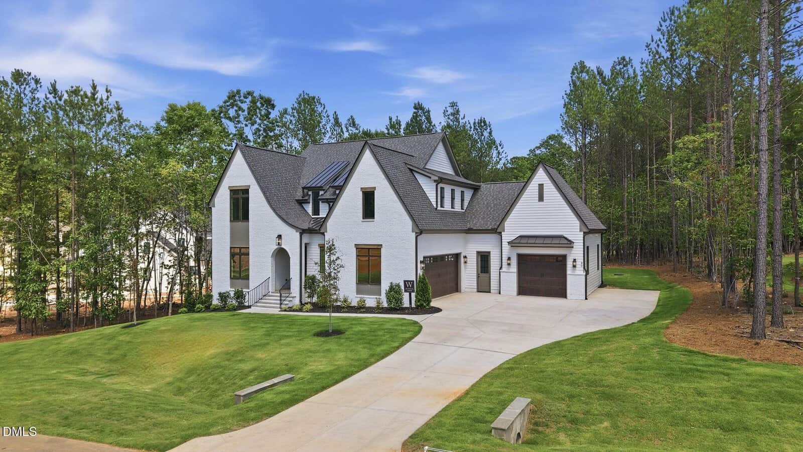 Modern two-story white house with dark roof, large driveway, three-car garage, manicured lawn, and surrounded by tall trees under a blue sky.