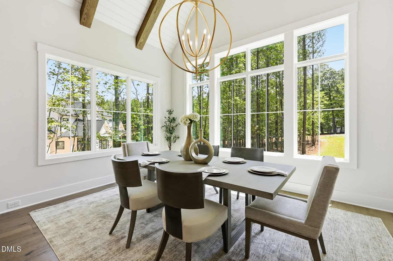 Modern dining room with a rectangular table set for six, large windows showing a wooded view, light walls, exposed wood beams on the ceiling, and a contemporary chandelier above the table.