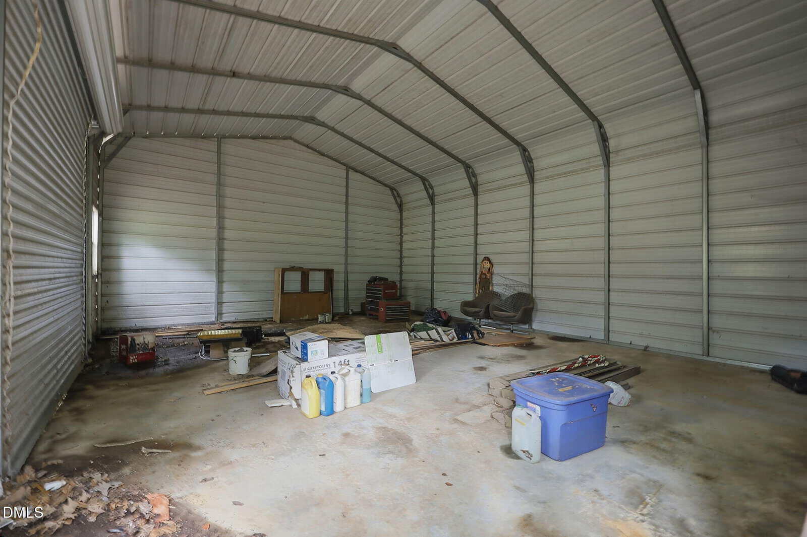 Interior of a large metal shed at 9531 Rueben Hart Road, Oxford, with a concrete floor, scattered tools, containers, and various items, including a blue cooler, jugs, boards, and a tire against the corrugated metal walls.