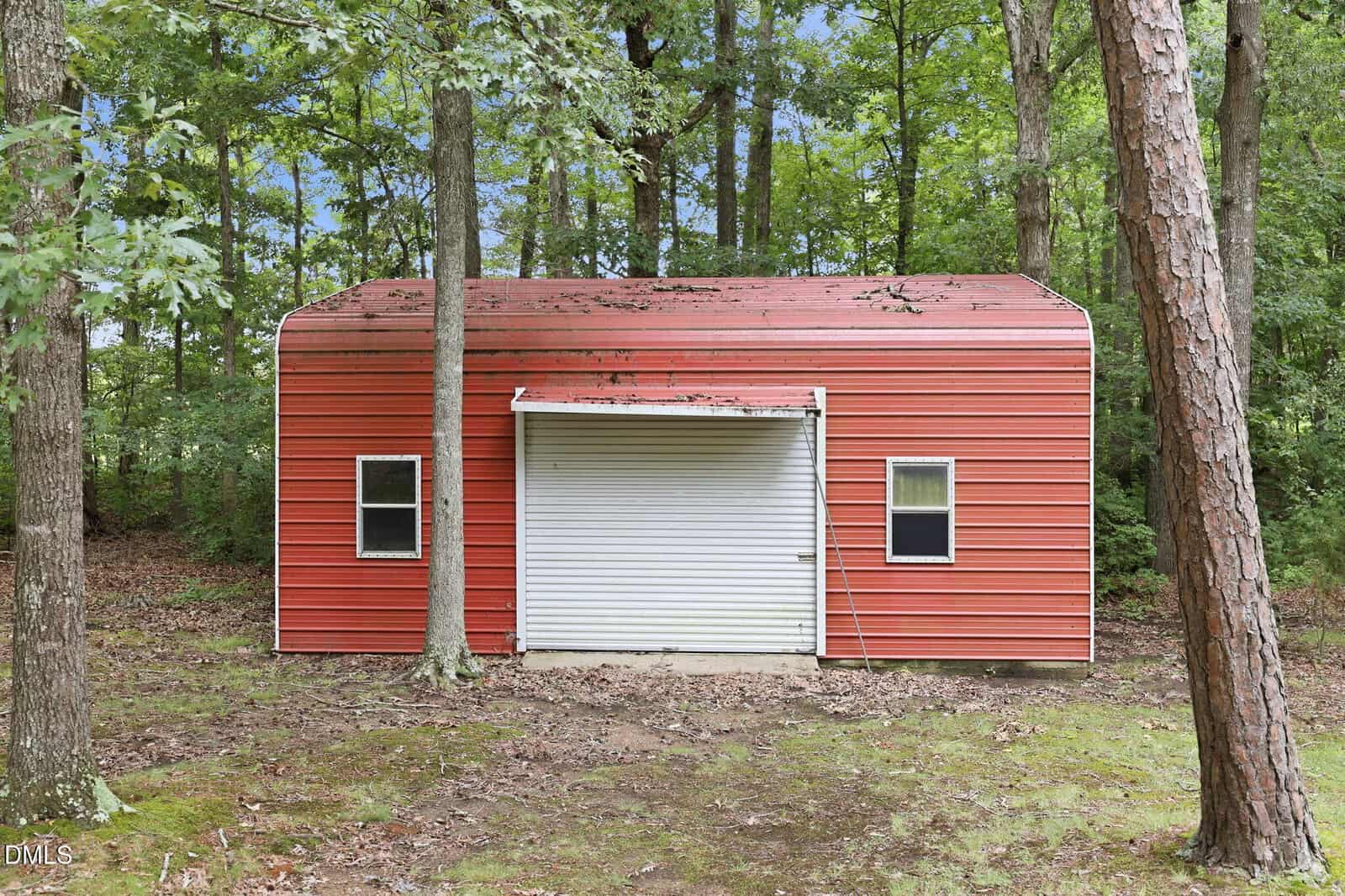A red metal shed with a white roll-up door and two small windows stands in a wooded area surrounded by trees and grass.