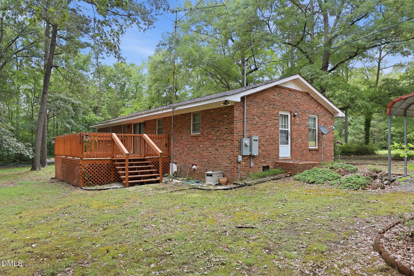 A red brick one-story house with a wooden deck and steps, surrounded by trees and a grassy yard. Utility meters and cables are visible on the side of the house.