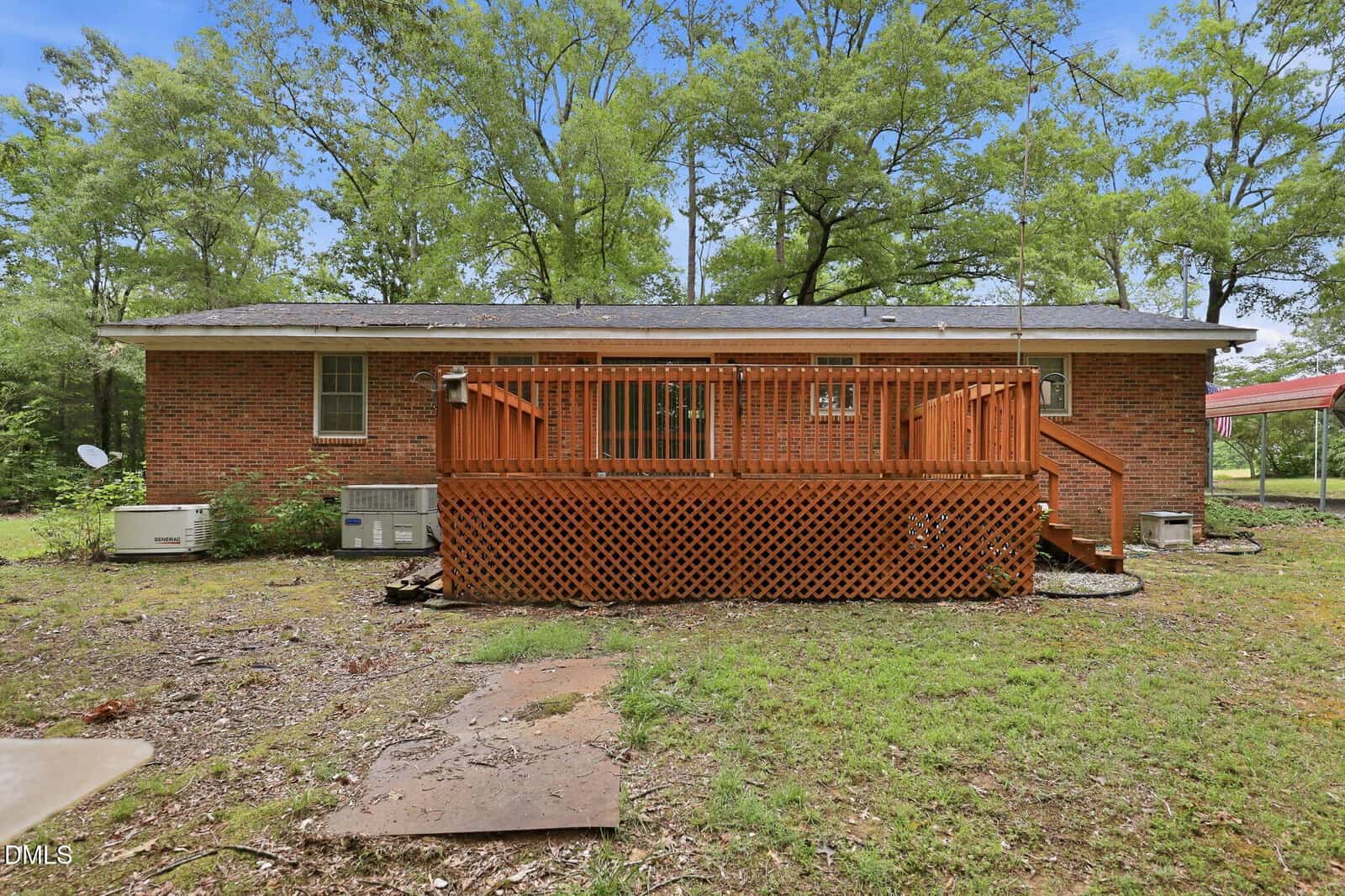A brick house with a raised wooden deck in the backyard, surrounded by grass, trees, and outdoor HVAC units. The sky is partly cloudy and the area appears peaceful and rural.