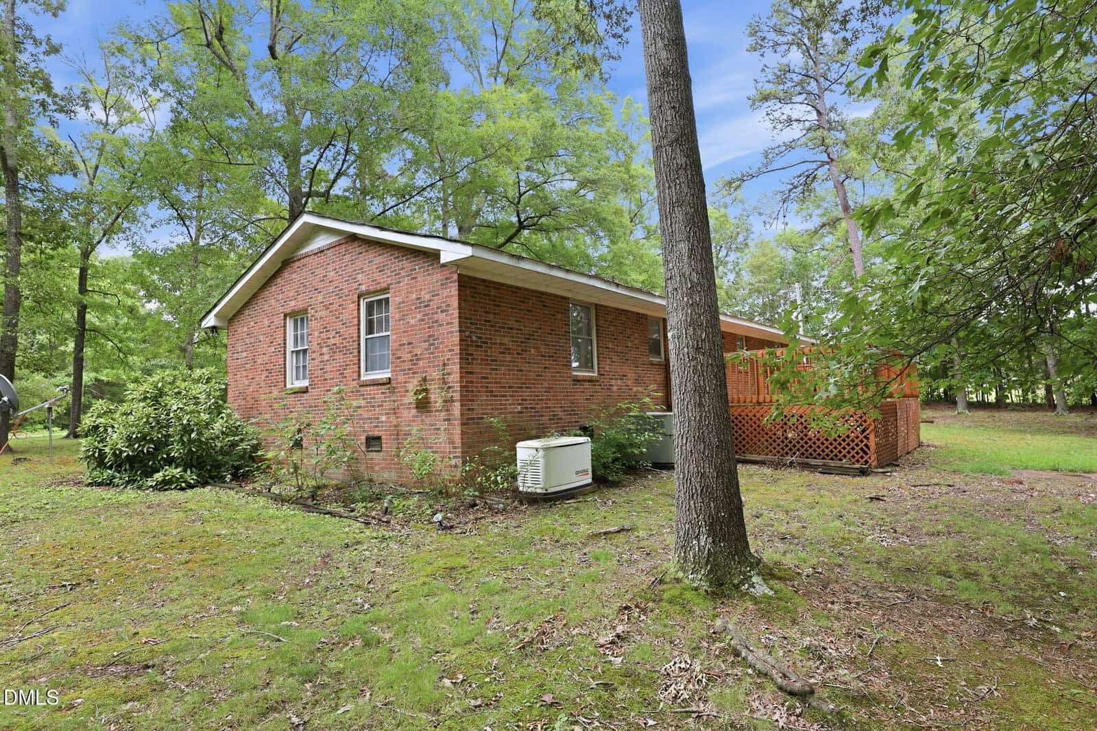 A brick house with white trim sits among tall trees and greenery. A wooden deck extends from one side, and an air conditioning unit is installed outside. The yard is grassy with patches of dirt and shaded by trees.