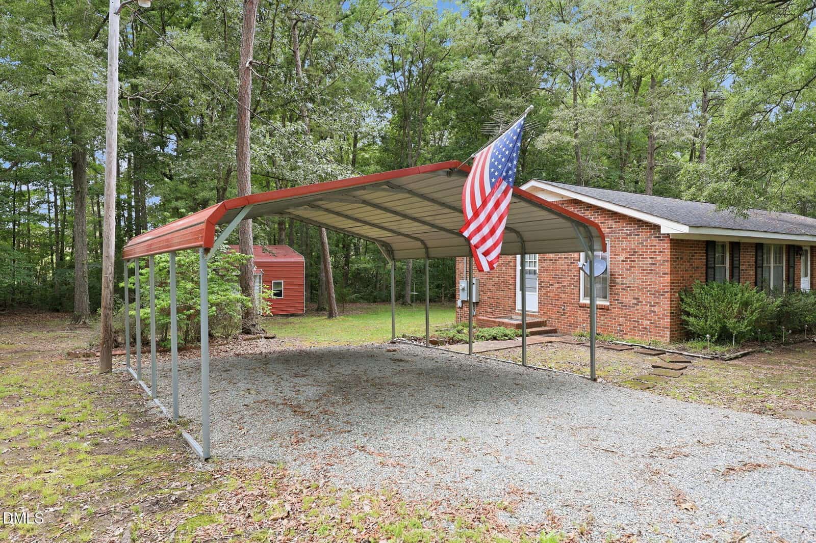 A metal carport with a gravel driveway and an American flag stands in front of a brick house surrounded by tall trees and greenery. A small red shed is visible in the background.