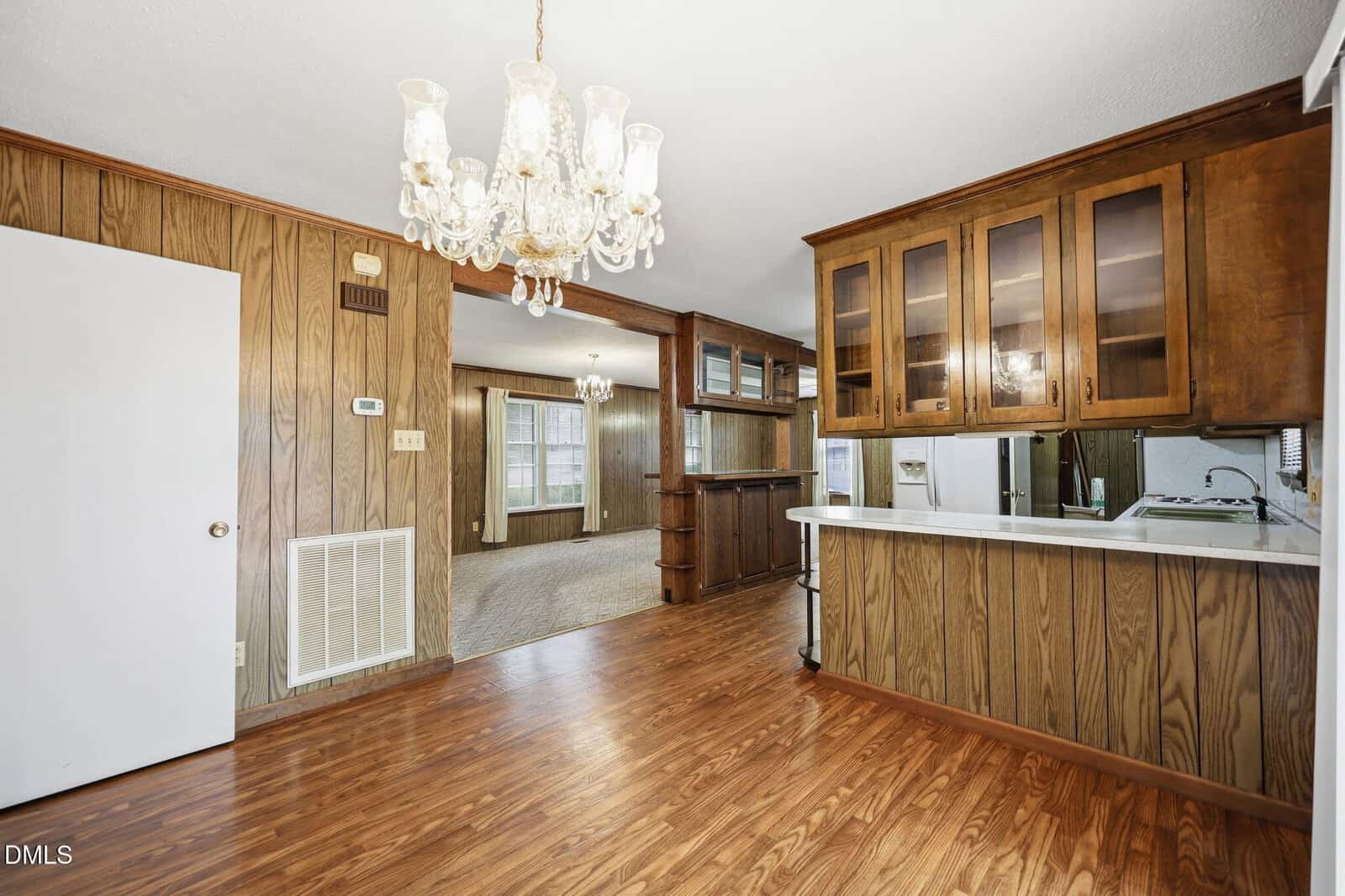 Dining area with wood-paneled walls, a chandelier, and wood flooring. Open view to living room and kitchen with glass-front cabinets and a breakfast bar. Bright natural light fills the rooms.