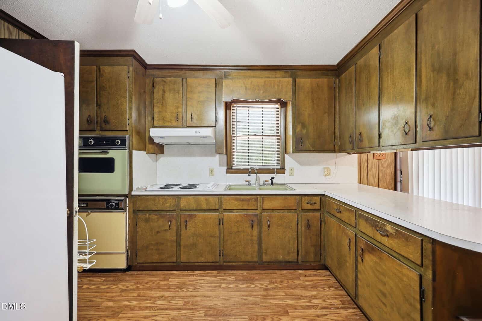 Vintage kitchen with greenish wood cabinets, white countertops, electric stovetop, old oven, and a window above the sink. Light wood flooring and ceiling fan visible.