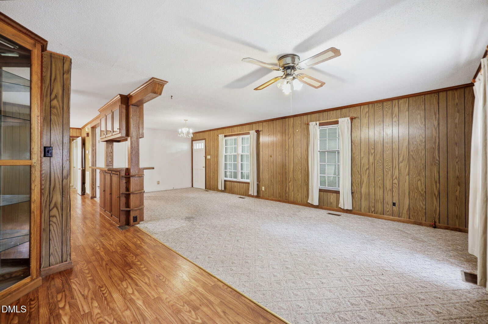 Spacious living room with wood-paneled walls, carpeted and wood flooring, ceiling fan, large windows with curtains, and an open dining area with a hanging light fixture.