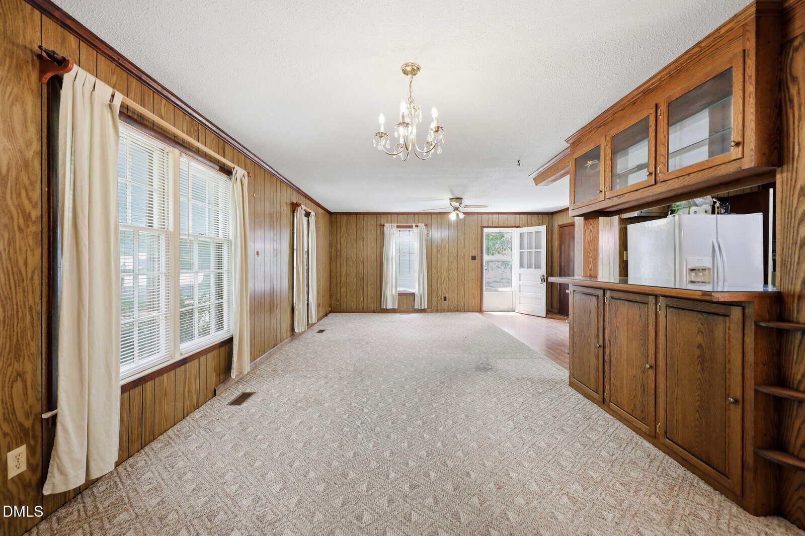 Spacious living room with wood-paneled walls, large windows, carpeted floor, chandelier, ceiling fan, and built-in wooden cabinets; a door leads to an adjacent kitchen with a visible refrigerator.