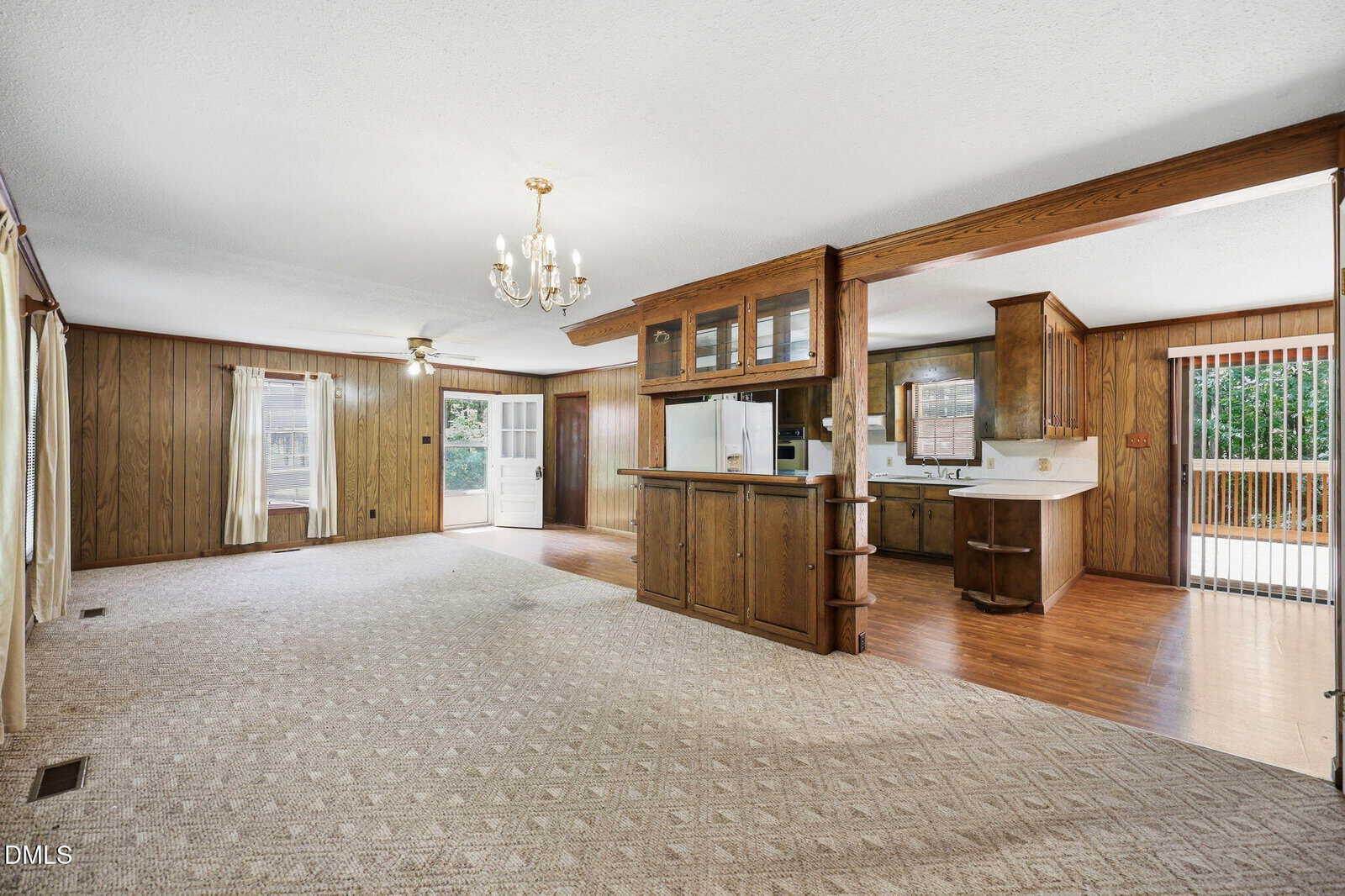 Spacious living and kitchen area with wood-paneled walls, carpet, hardwood floors, large windows, sliding glass door, chandelier, and built-in cabinetry separating the two spaces.