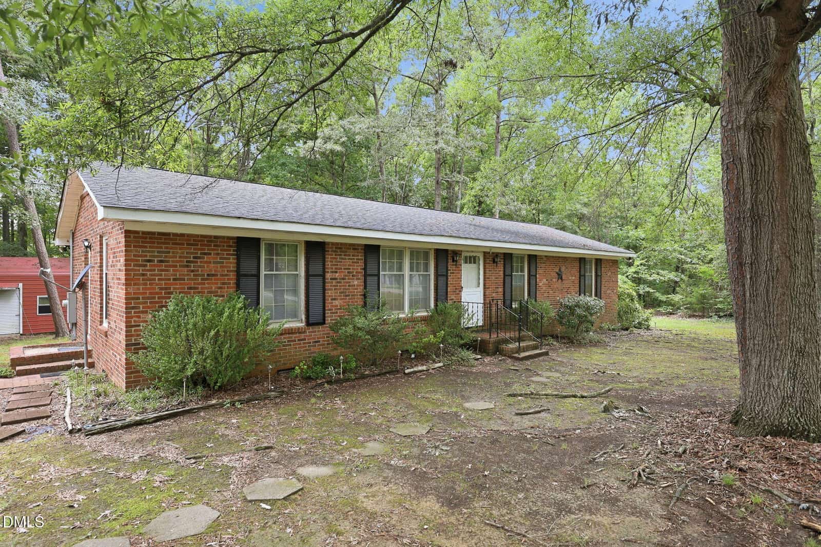 A single-story brick house with black shutters and a gray shingle roof sits surrounded by trees at 9531 Rueben Hart Road, Oxford. A small front porch with railings leads to the white front door, while stepping stones cross the mostly bare yard.