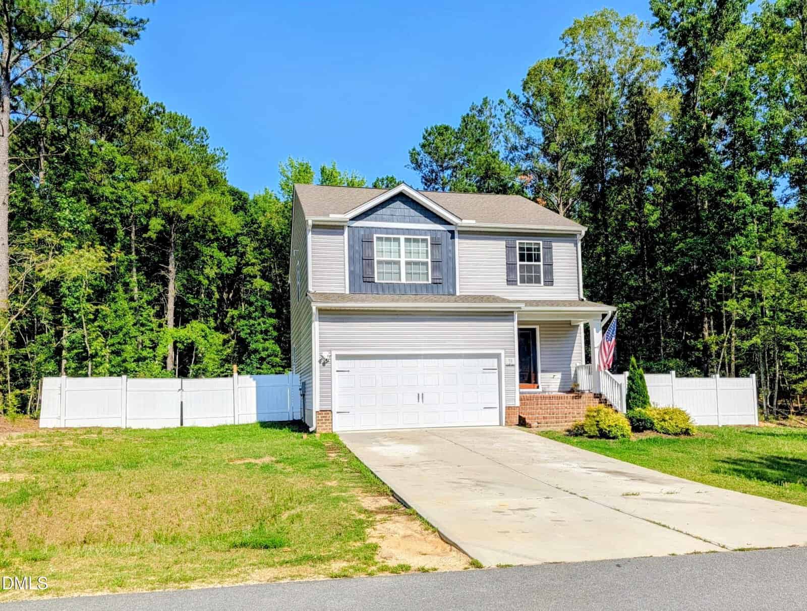 A two-story gray house with white trim, a double garage, and an American flag near the front porch. The house is surrounded by a white fence and trees, with a driveway and lawn in front.