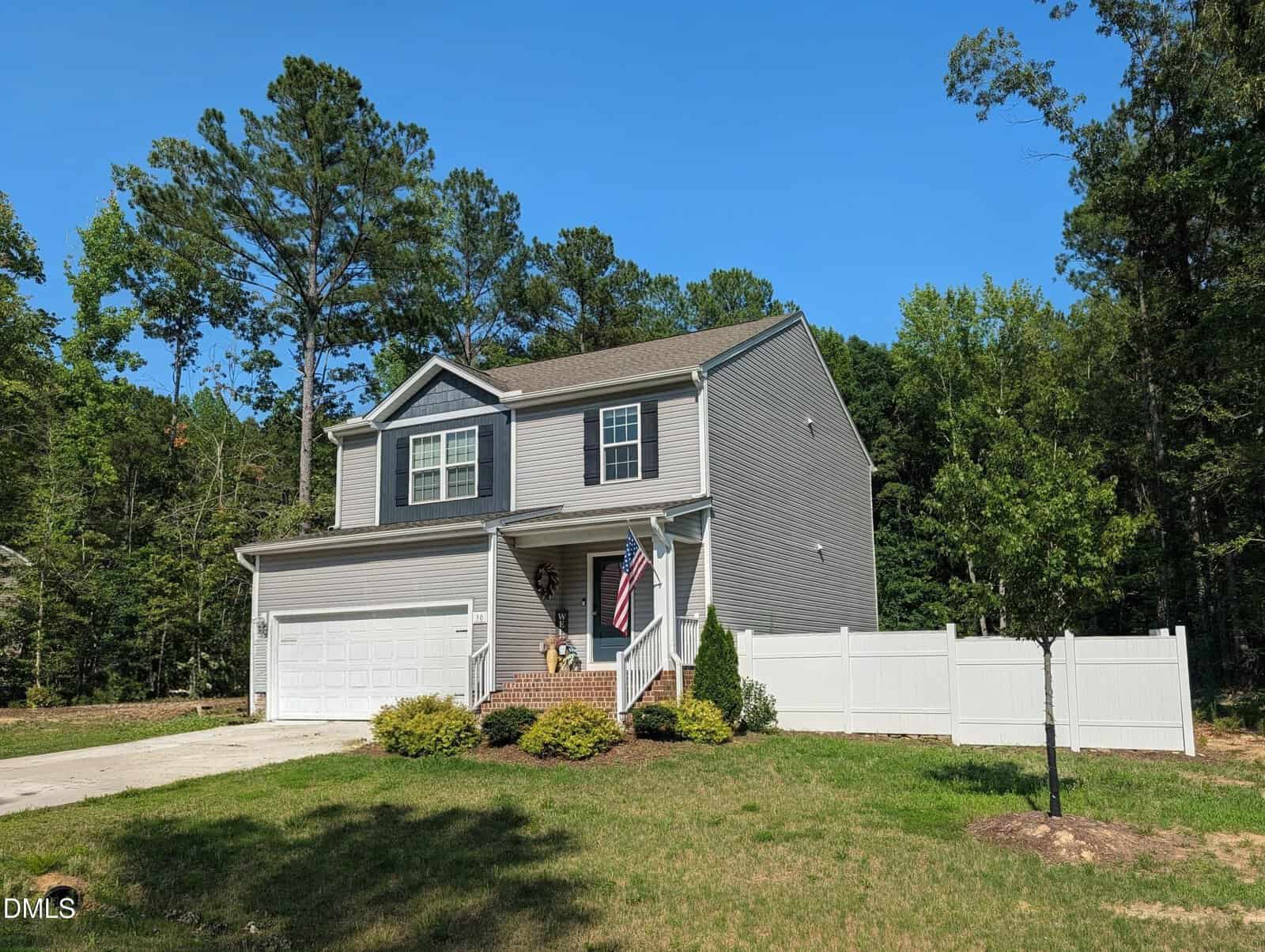 Two-story gray suburban house with white trim, a double garage, and an American flag by the entrance. The yard has green grass, shrubs, and is bordered by a white fence and tall trees in the background.