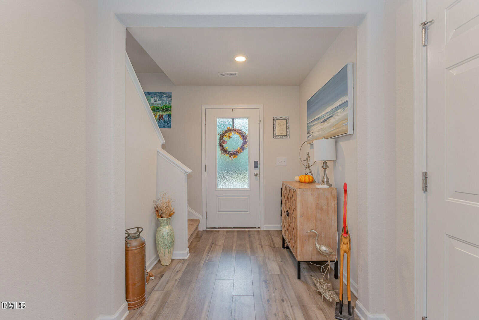 A bright hallway with wood flooring, a white door with a wreath, a staircase, coastal-themed art, a small cabinet, fall decorations, and an umbrella stand with a broom and walking stick.
