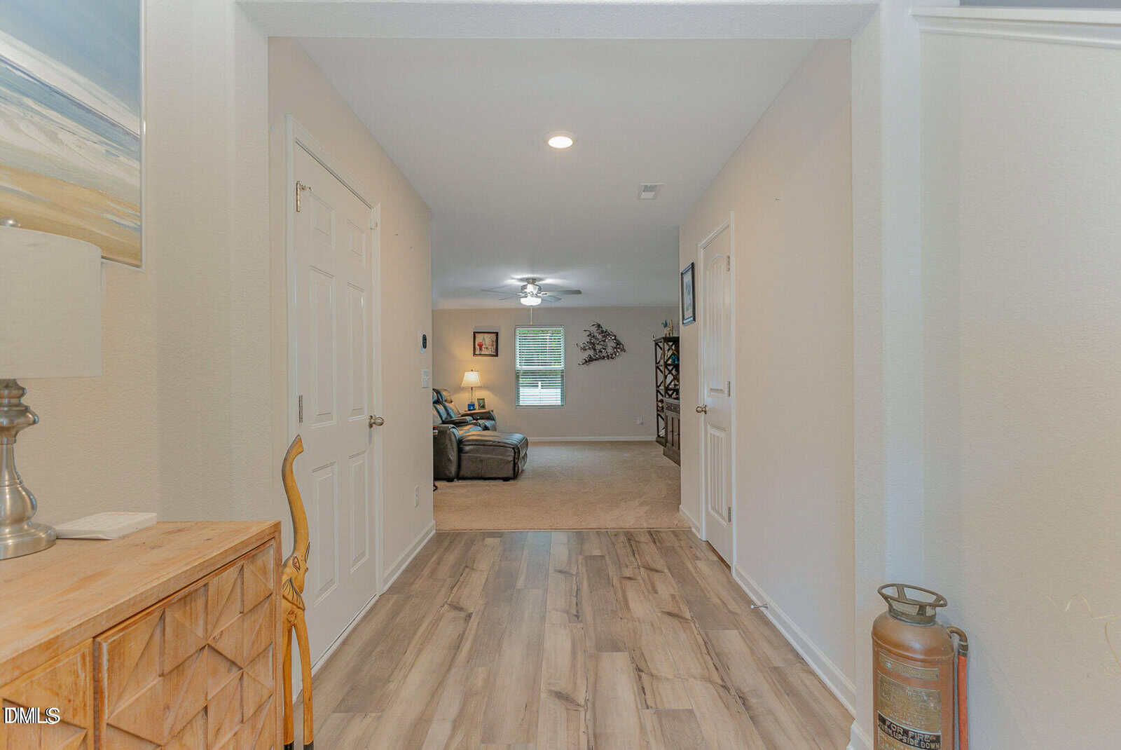 View of a hallway with light wood floors, a wooden cabinet with a lamp, a walking cane, and a fire extinguisher. The hallway leads to a well-lit living room with furniture and wall art visible.