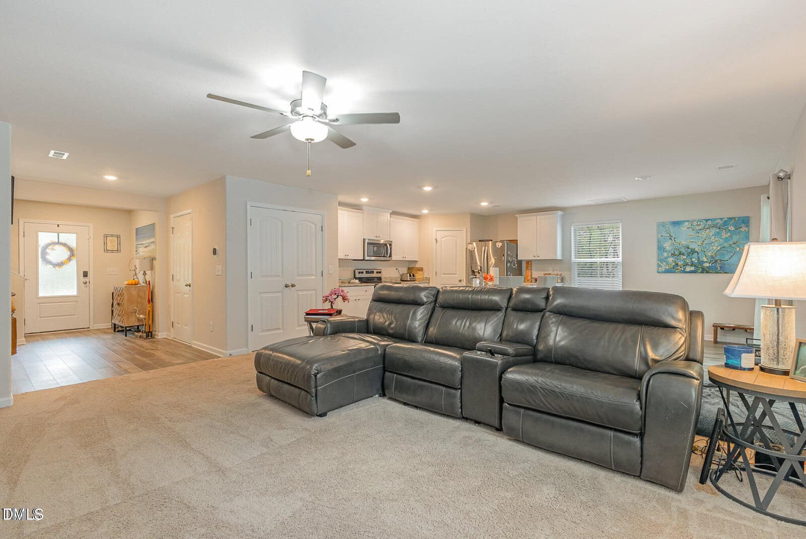 Open living room with a dark leather sectional sofa, beige carpet, and a ceiling fan, leading to a kitchen with stainless steel appliances. Natural light from windows, light walls, and recessed lighting brighten the space.