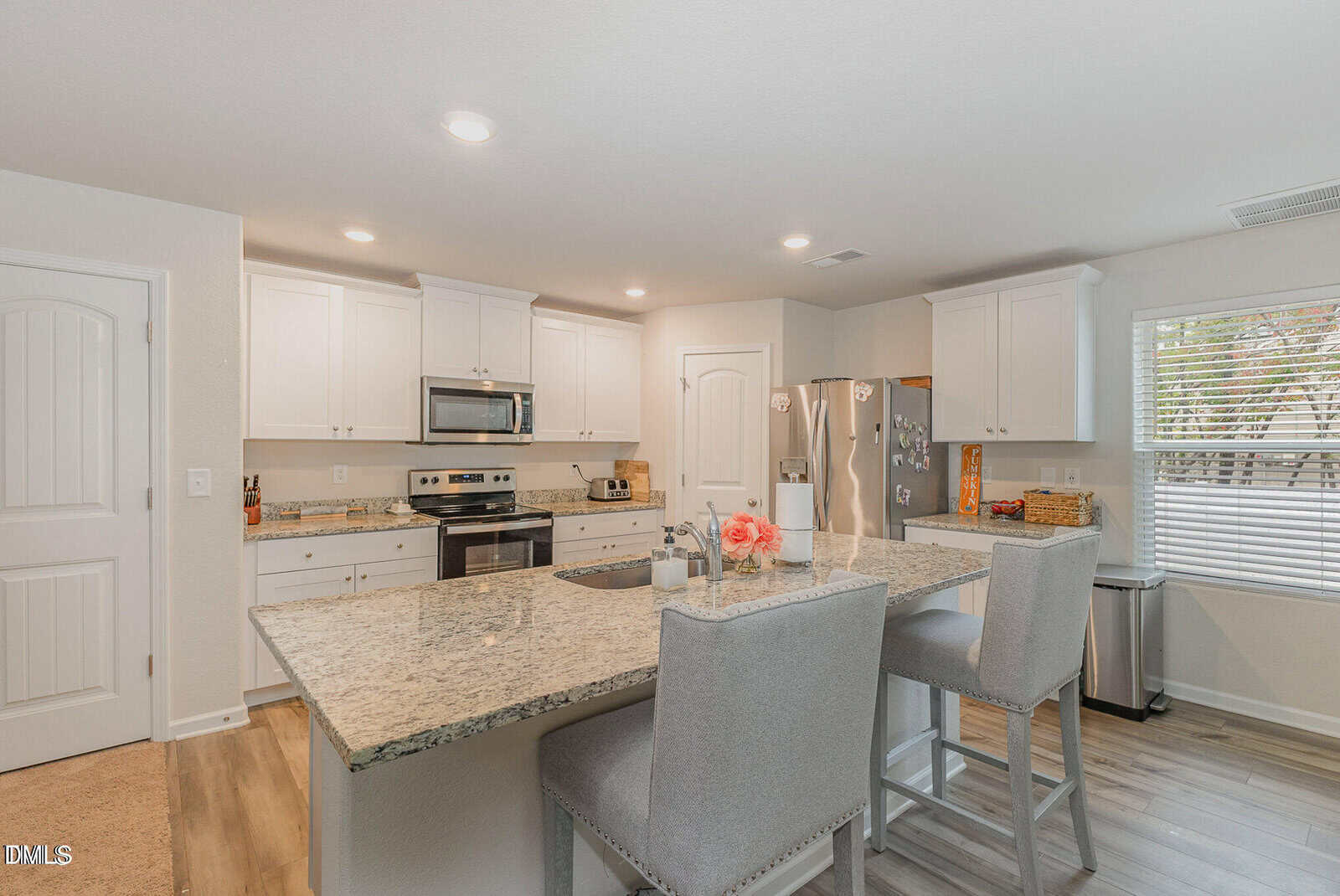 Modern kitchen with white cabinets, stainless steel appliances, granite countertops, a large island with a sink, two upholstered chairs, hardwood floors, and a window with blinds letting in natural light.