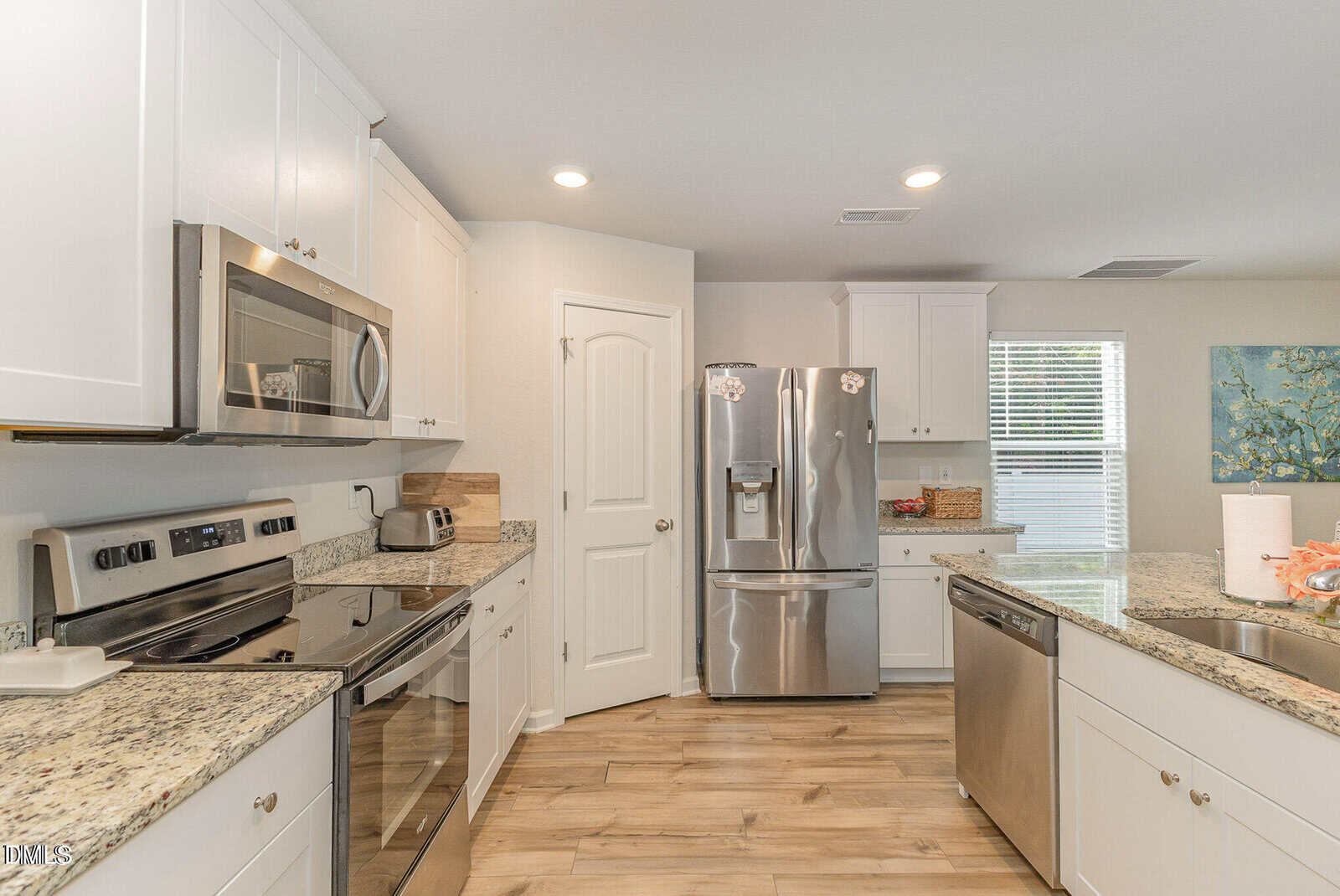 Modern kitchen with stainless steel appliances, granite countertops, white cabinets, hardwood floors, and an island with a sink. A window with blinds and a painting are visible in the background.