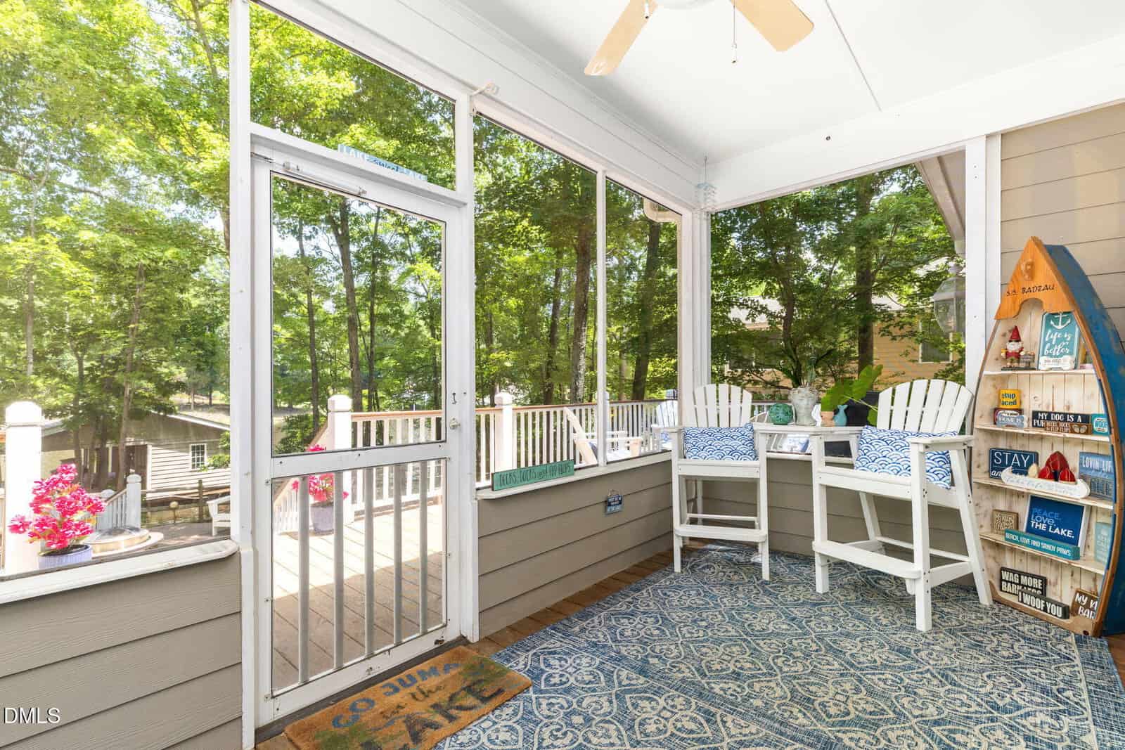A screened-in porch with white chairs, blue cushions, a patterned rug, and a small table. The porch overlooks a deck and trees, with decorative lake-themed signs on the wall and flowers on the railing.