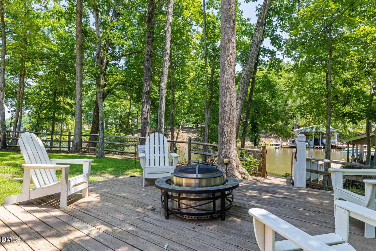 Wooden deck with four white Adirondack chairs surrounding a fire pit, shaded by tall trees, near a fence and a dock overlooking a calm, tree-lined lake.