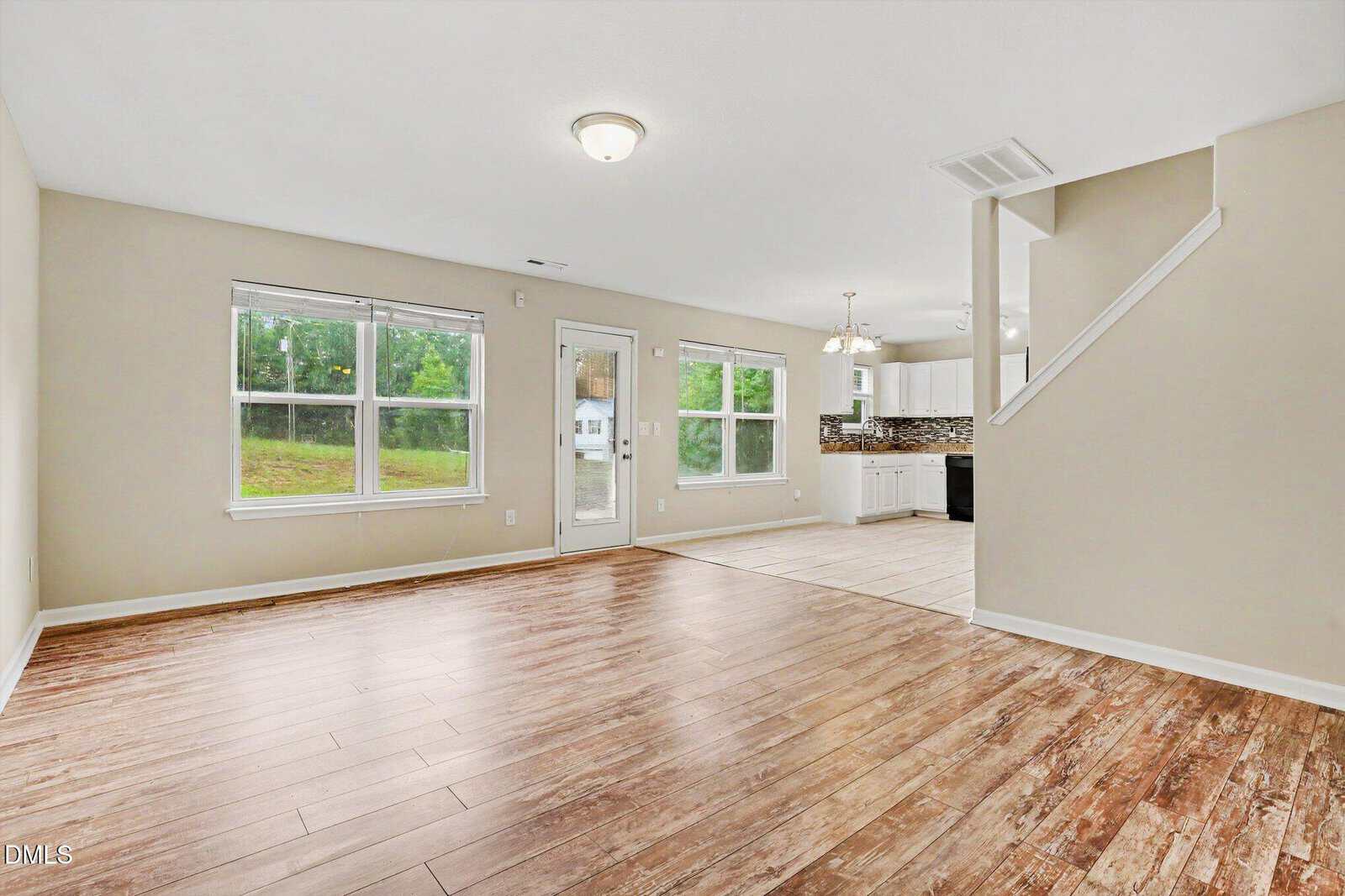 Spacious living area with wood-look flooring, beige walls, large windows, and adjacent dining area; kitchen with white cabinets and tiled floor visible in the background near stairs.