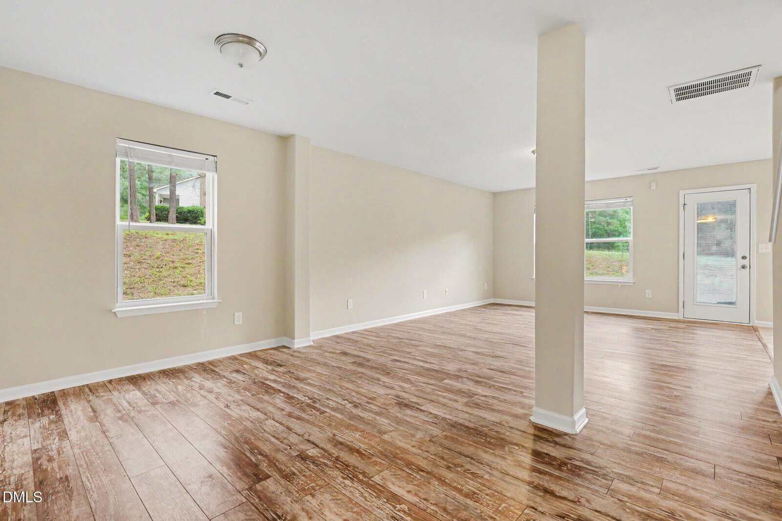 Spacious, empty room with light beige walls, wood-style flooring, two large windows, a glass door leading outside, and a support column in the center. Ample natural light enters the space.