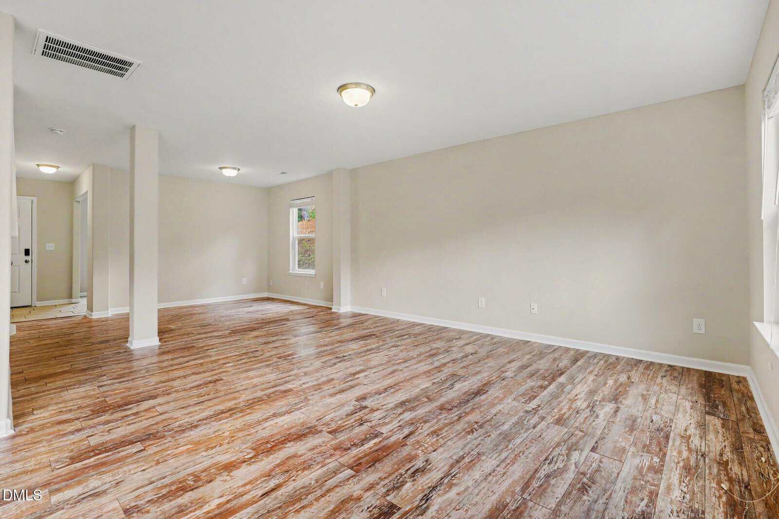 Spacious empty room with light beige walls, wood-patterned vinyl flooring, ceiling lights, and several windows letting in natural light. A hallway and white front door are visible in the background.