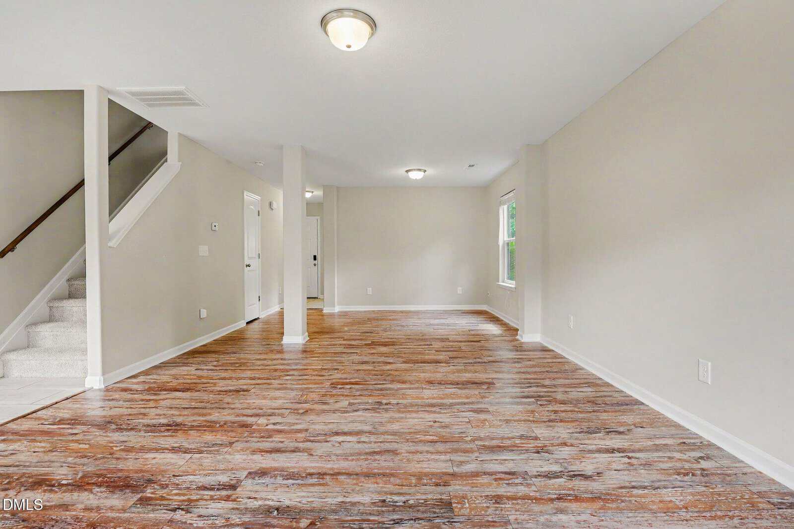 Open-concept living area with wood-look flooring, neutral walls, ceiling lights, a staircase to the left, a window to the right, and an entryway in the background.