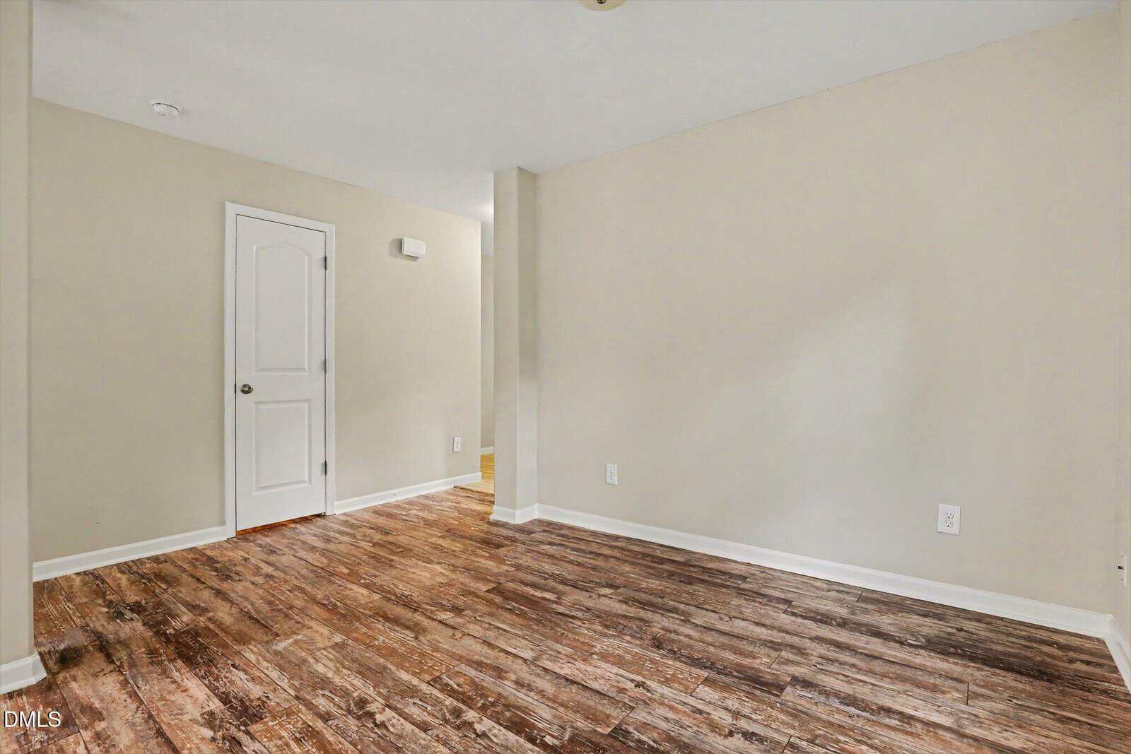 Empty room with light beige walls, white trim, and wood-patterned flooring. A closed white door and a small wall divide the space. Bright, modern, and clean with visible electrical outlets.