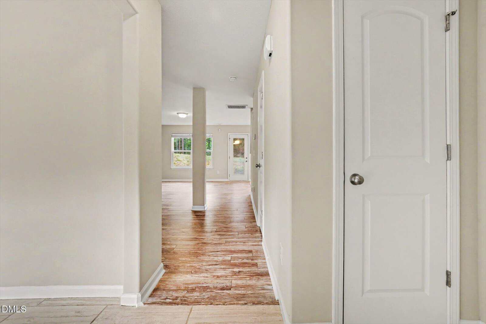 Bright hallway with light-colored walls and a closed white door on the right, leading to an open living area with large windows and wood flooring.