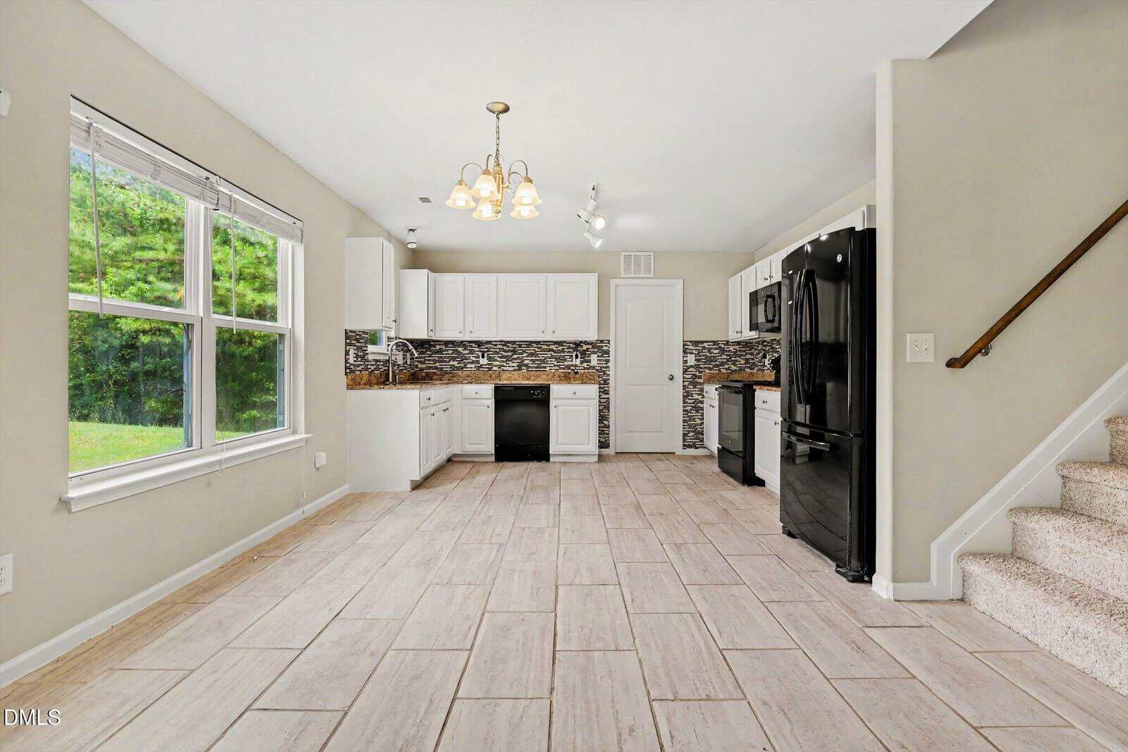 Modern kitchen with white cabinets, black appliances, tiled floors, mosaic backsplash, large windows with a view of greenery, and carpeted stairs leading up on the right.