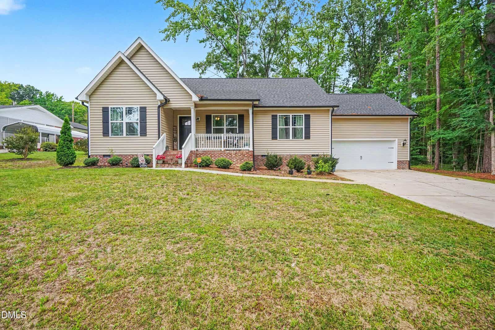Charming single-story beige house at 212 Tanglewood Drive, Louisburg, featuring a covered front porch with white railing and attached garage. Surrounded by grass, shrubs, and trees, with a concrete driveway on the right.