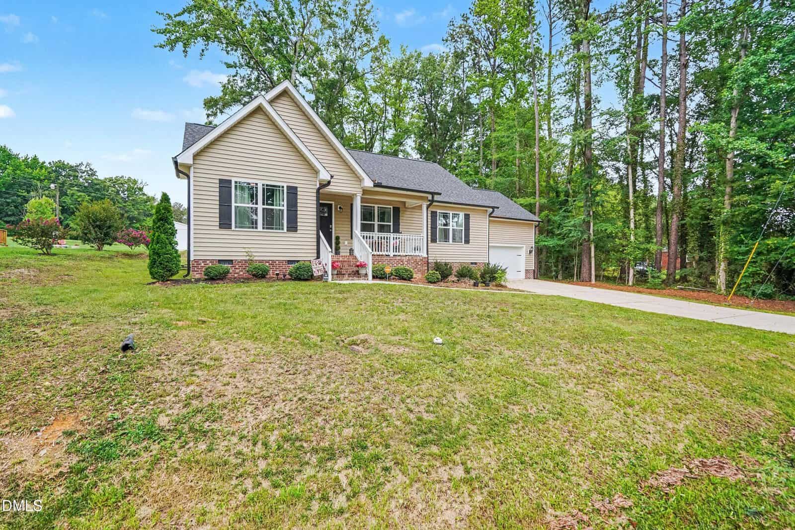 Single-story beige house with white trim, front porch, and attached garage, set on a spacious lawn with surrounding trees and a long driveway on the right side.