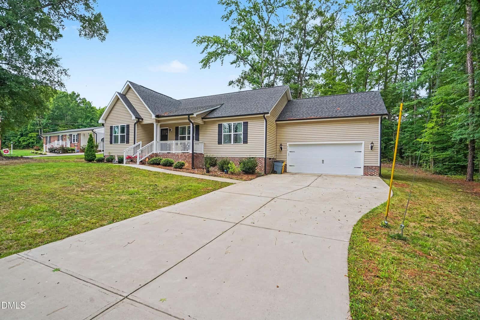 Single-story beige house with a covered front porch, white trim, and attached two-car garage. A wide concrete driveway leads to the garage, and the home is surrounded by grass, shrubs, and tall trees.