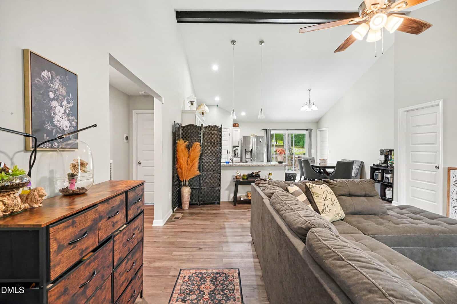 Modern open-concept living space with a gray sectional sofa, kitchen island, dining area, and ceiling fan. Decor includes plants, a wooden dresser, and a room divider, with wood flooring throughout.