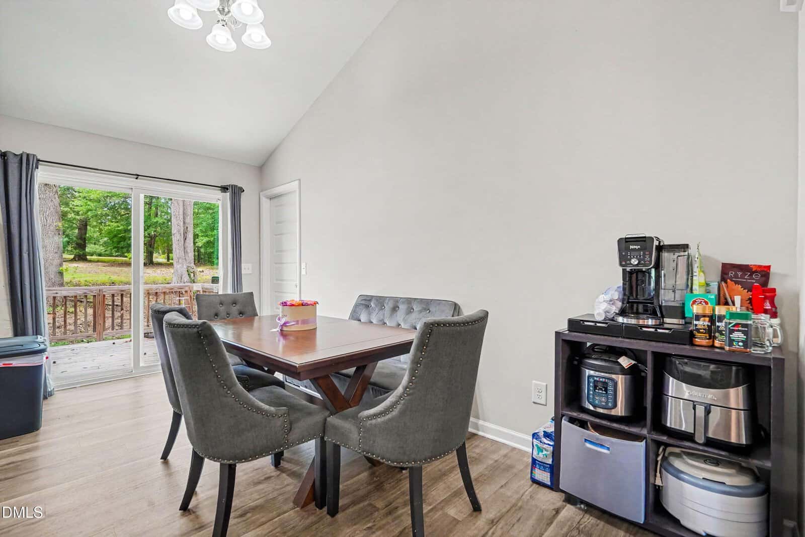 Dining area with a wooden table, four gray tufted chairs, and a beverage station with a coffee maker and small appliances. Sliding glass doors open to a deck with a wooded view.