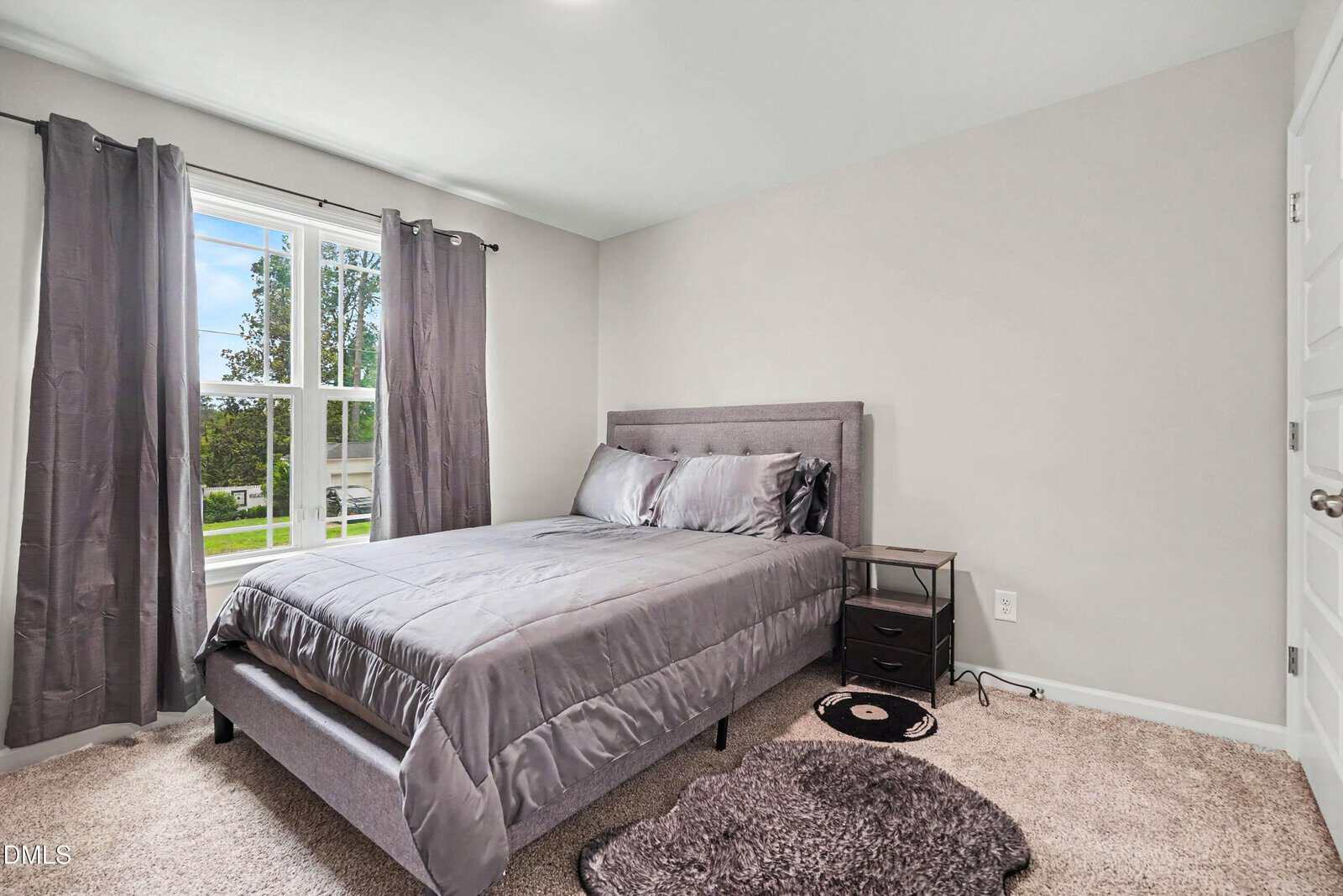 Bedroom with a gray bed, matching bedding, two gray curtains on a large window, a black nightstand with a lamp, and a small rug on beige carpet. Walls are light gray and the room is well lit.