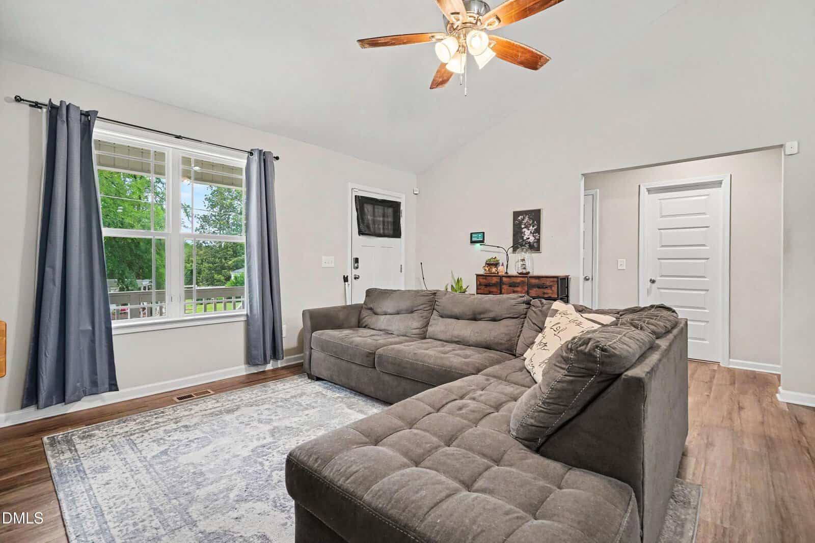 Bright living room with a large gray sectional sofa, patterned rug, ceiling fan with lights, wooden floors, and a window with dark curtains overlooking greenery outside. A door and dresser are also visible.