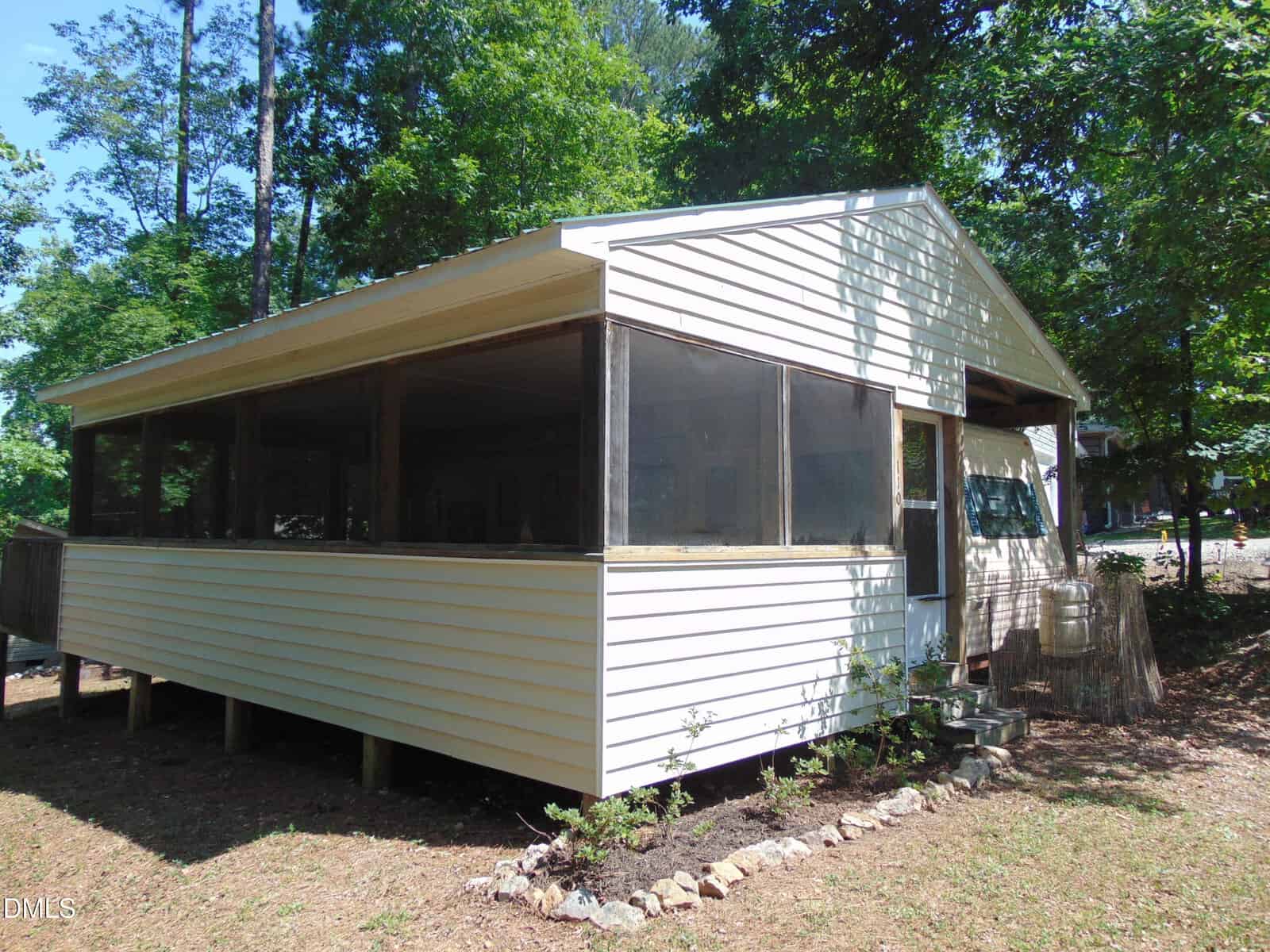 A small, white, single-story building with a large screened porch, surrounded by trees and plants, sits on a plot of dirt and grass. The porch is elevated, and sunlight filters through the trees above.