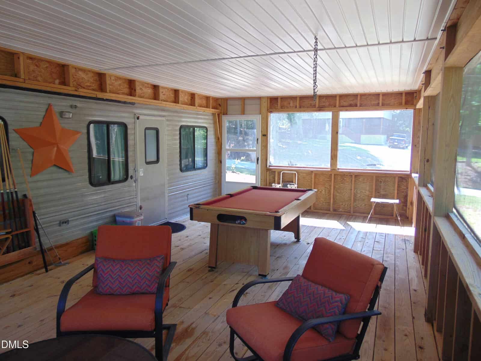 A screened-in porch with wood floors and ceiling features a pool table, round table with orange cushioned chairs, cues in the corner, and a large orange star mounted on the side of a trailer. Sunlight streams through the windows.