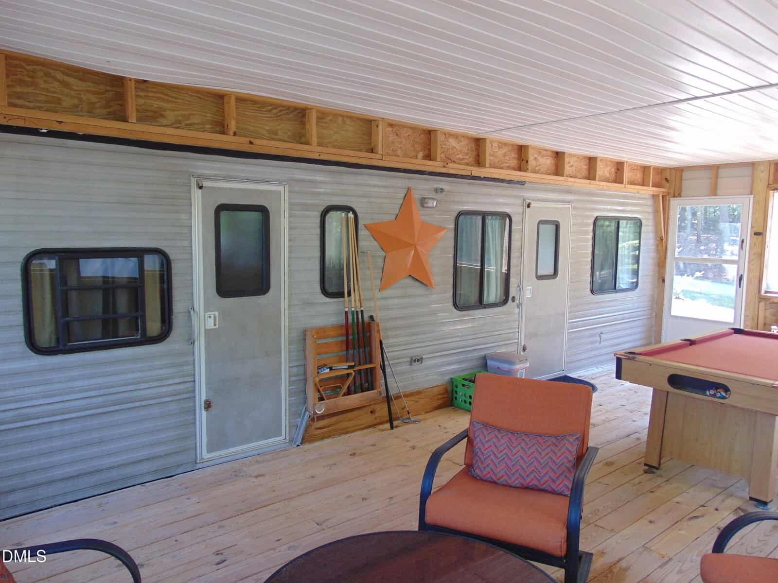 A covered porch area with wood flooring features the side of a trailer, decorated with a large orange star. There are patio chairs, a table, and a pool table present, with trees visible outside the windows.