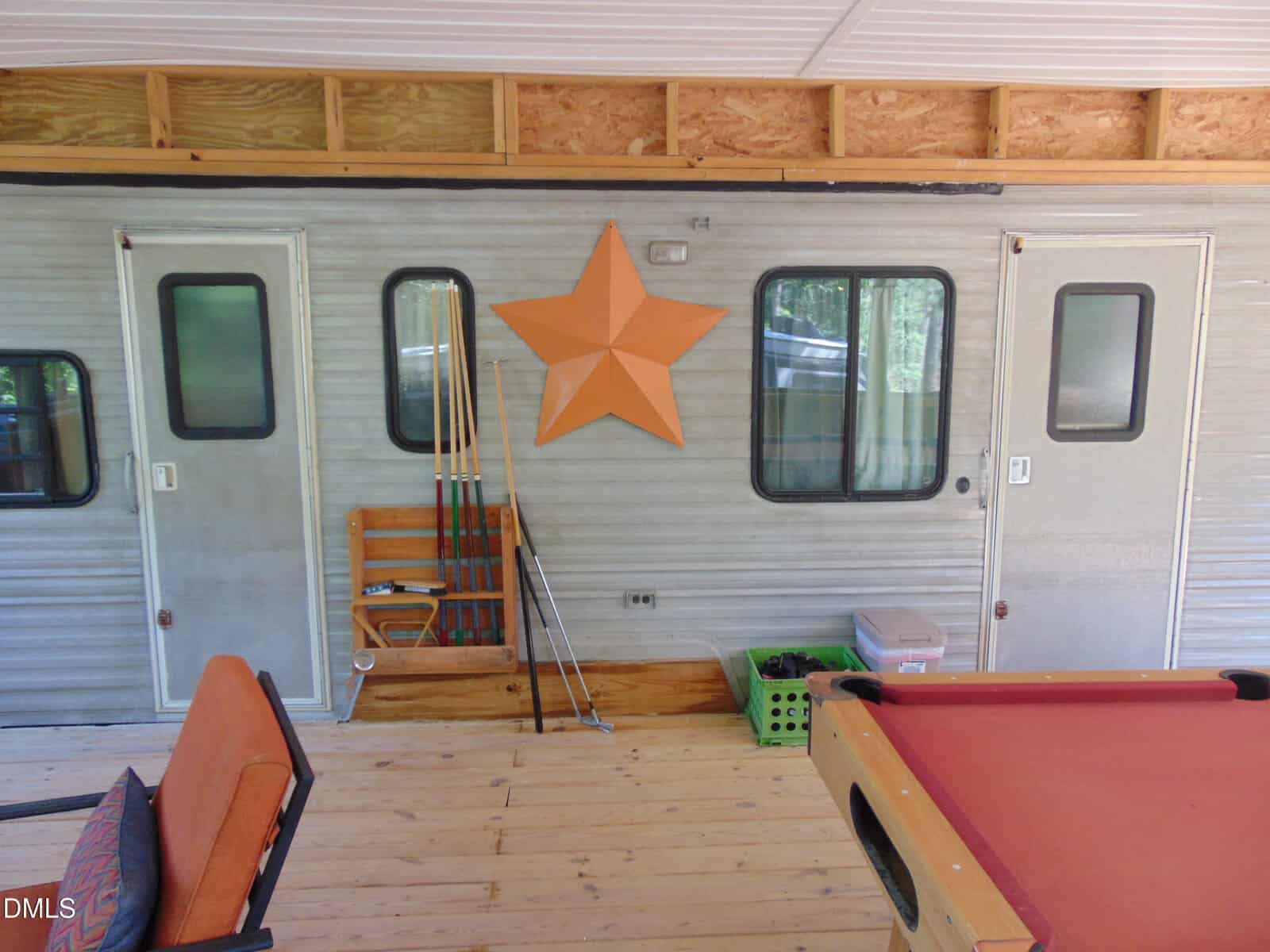 A large orange star hangs on a trailer wall between two doors. Pool cues are stored in a rack, and a red pool table and orange chairs are on the wooden deck.