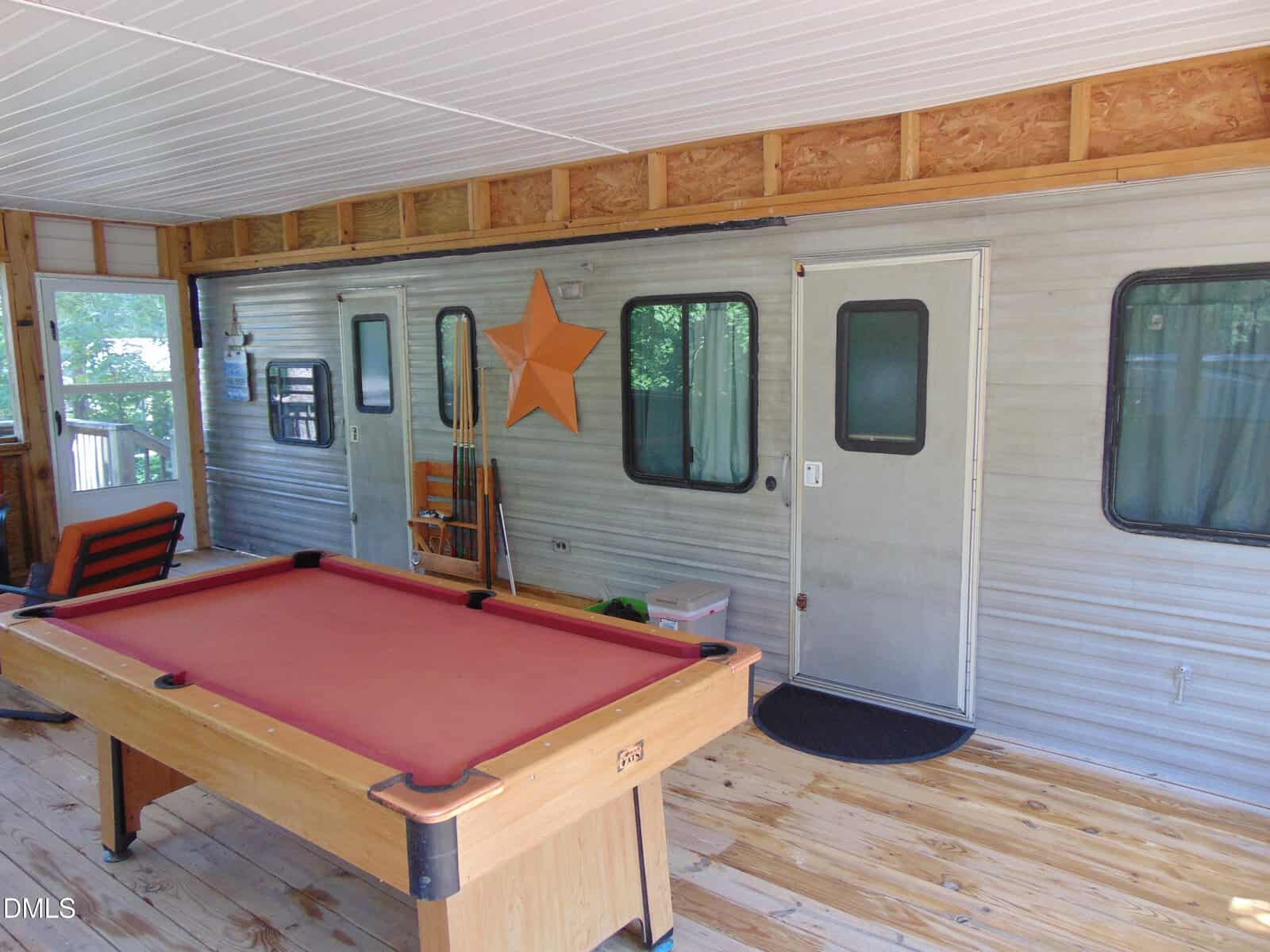 A red felt pool table sits on a wooden floor in front of a silver trailer wall with two doors, a star decoration, pool cues, and windows.