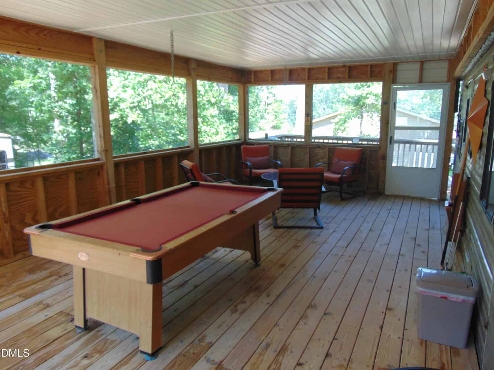 Screened porch with wooden floor, a red pool table, four orange cushioned chairs around a round table, and a door leading outside. Large windows let in natural light and show green trees outside.
